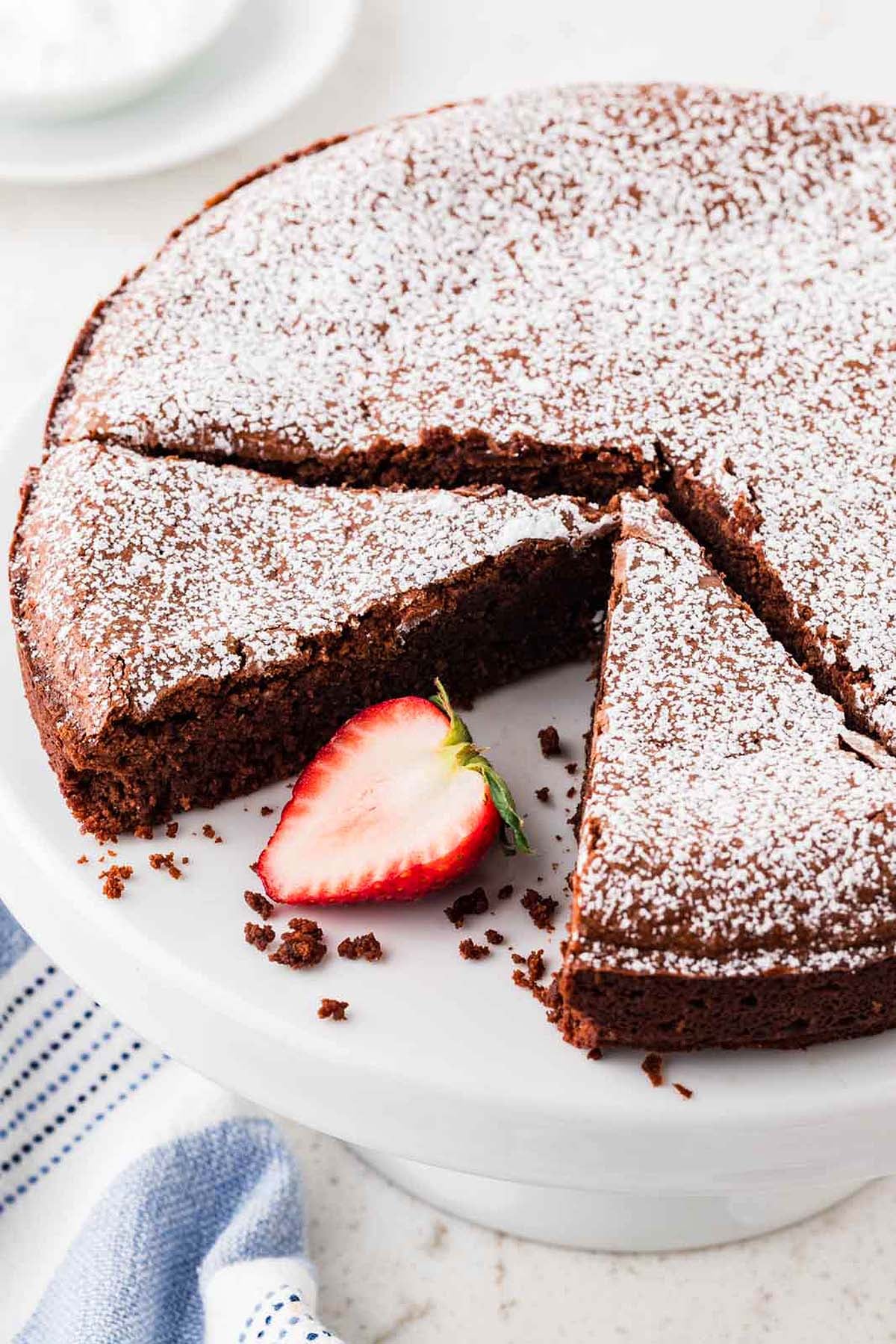 Chocolate torta caprese dusted with powdered sugar and a single strawberry slice on a white cake stand.