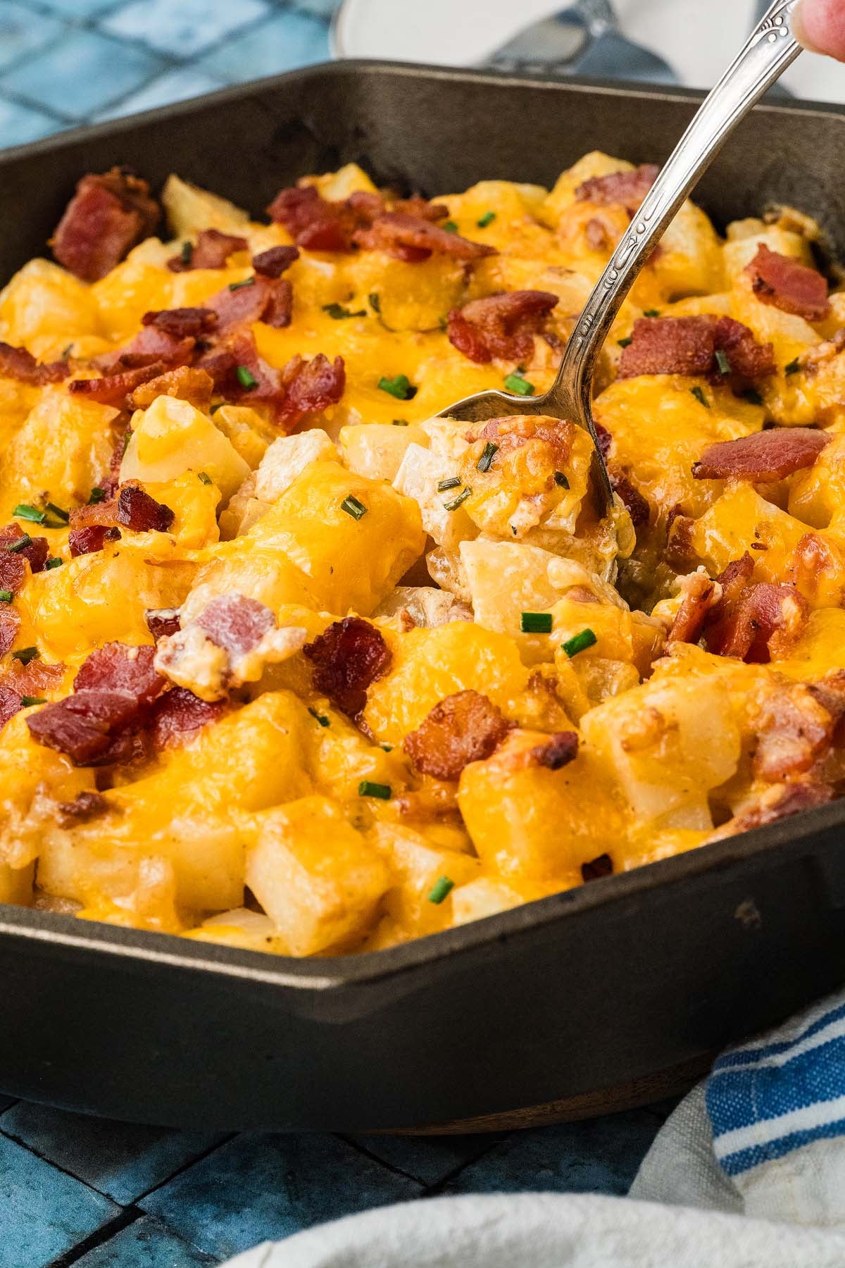 Skillet of cheesy potatoes with bacon and chives being served with a spoon.
