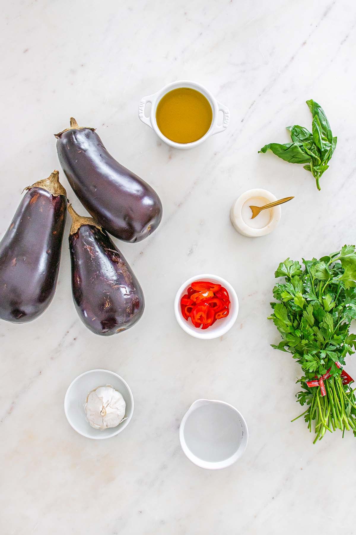 Three eggplants, fresh herbs, garlic, sliced red chili, and small bowls of oil and water on a marble surface.