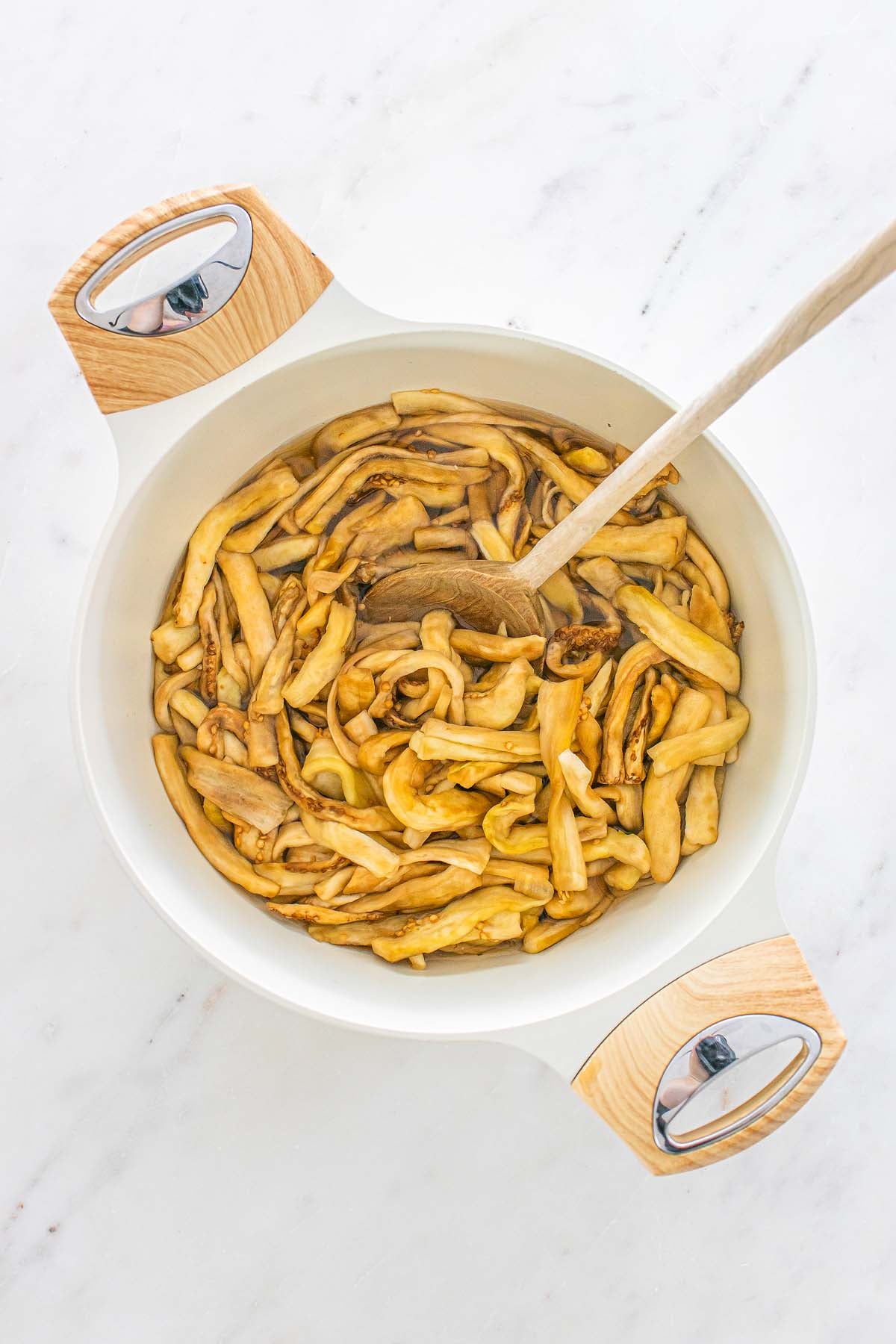 Strips of cooked eggplant in a white pot with a wooden spoon.