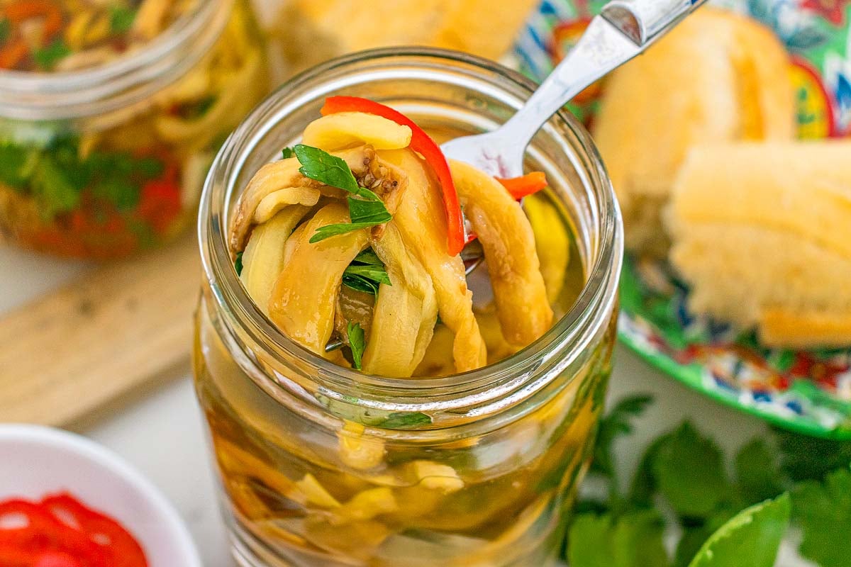 A fork lifting pickled eggplant strips with herbs and red pepper from a glass jar.