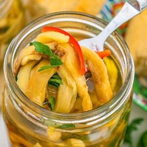 A fork lifting pickled eggplant strips with herbs and red pepper from a glass jar.
