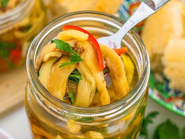 A fork lifting pickled eggplant strips with herbs and red pepper from a glass jar.