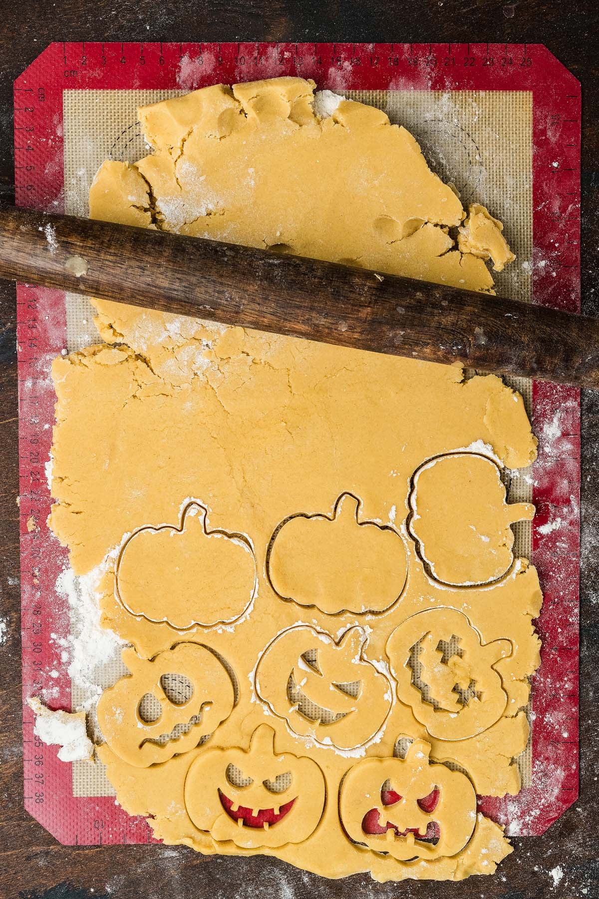 A rolling pin rests on dough being cut into pumpkin and jack-o’-lantern shapes on a baking mat.