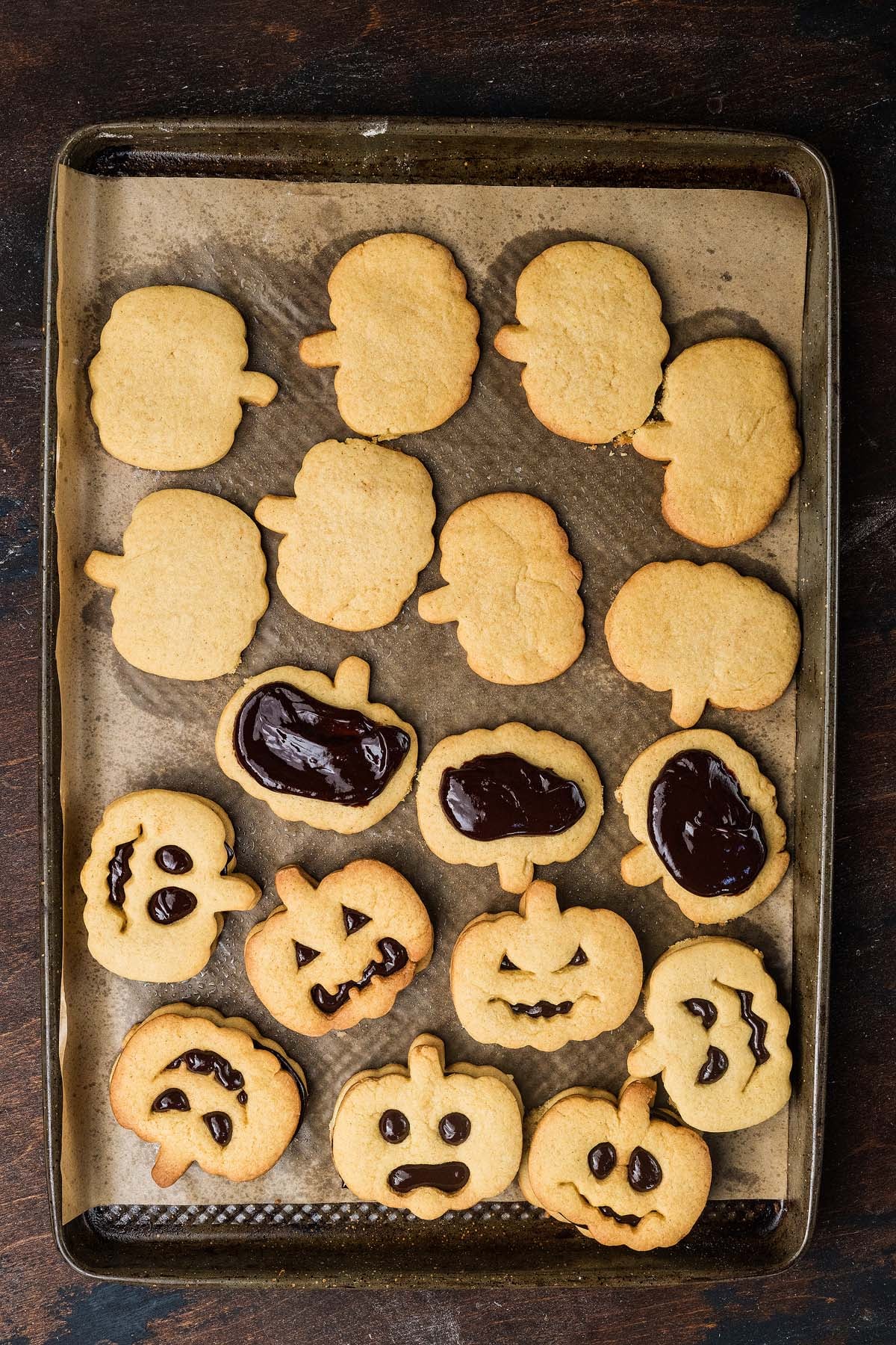 Pumpkin-shaped cookies on a baking sheet, some plain and some decorated with chocolate jack-o’-lantern faces.