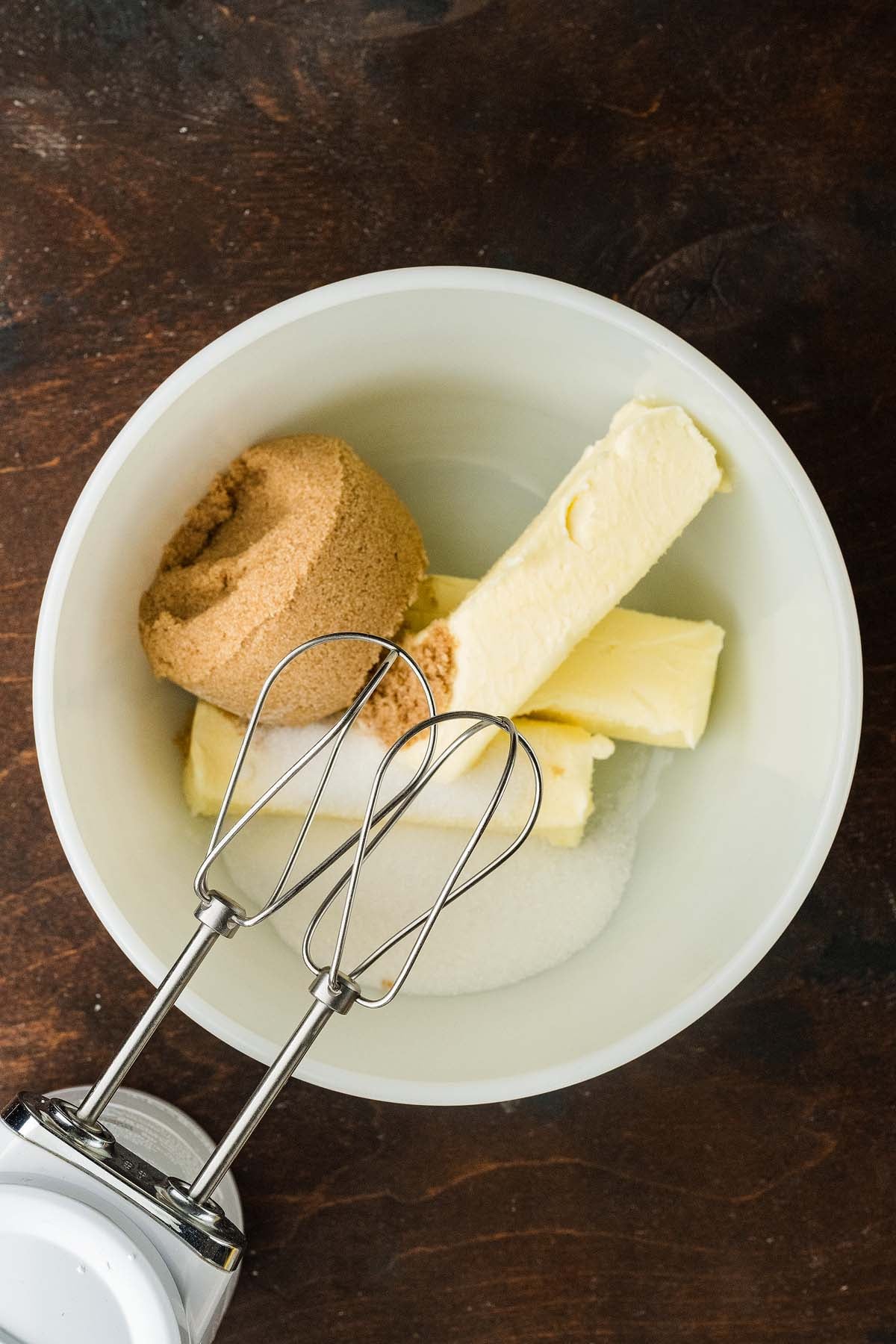 Mixing bowl containing sticks of butter, brown sugar, and white sugar with hand mixer beaters.