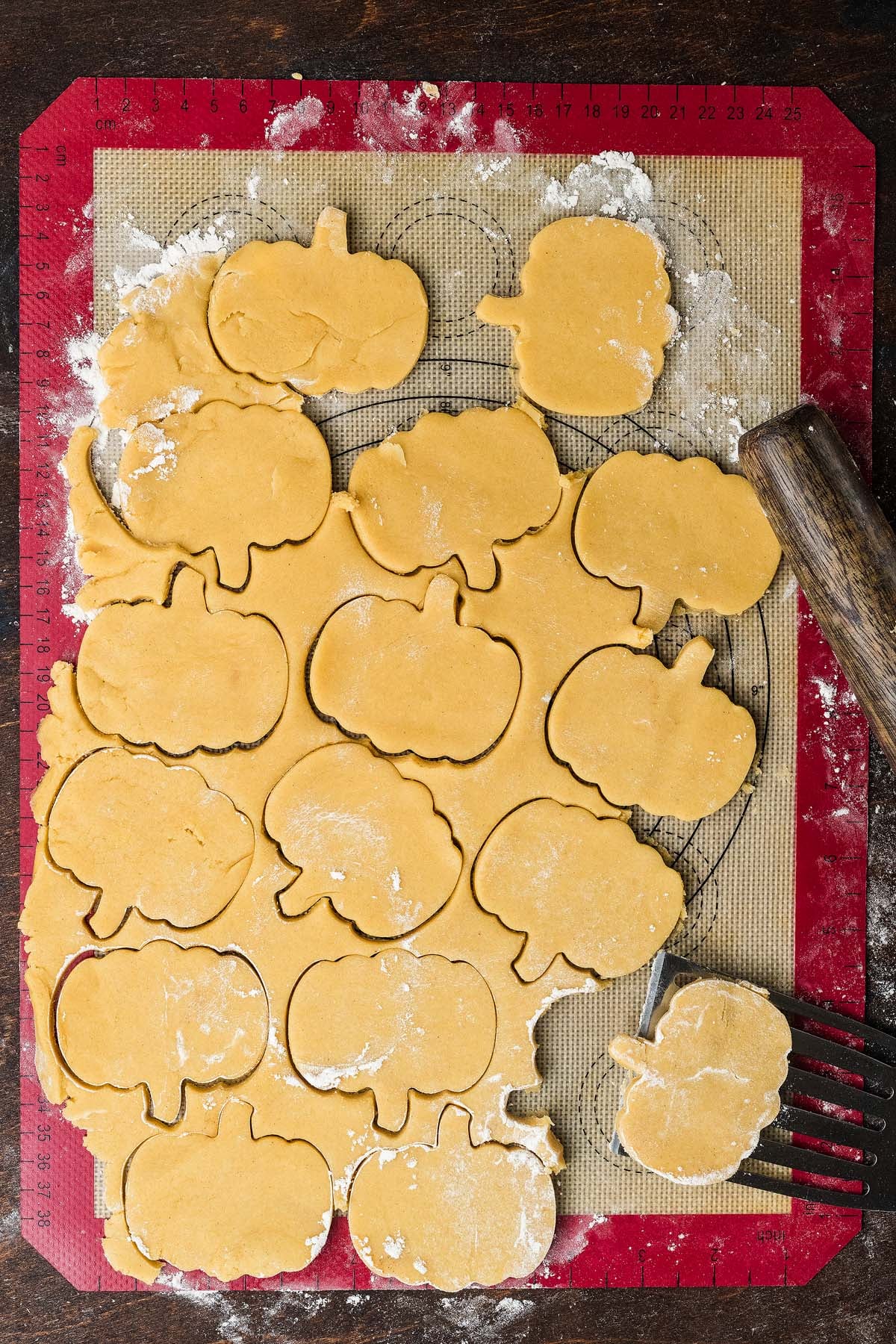 Dough rolled out on a silicone mat with several pumpkin-shaped cookies cut out and a spatula lifting one.