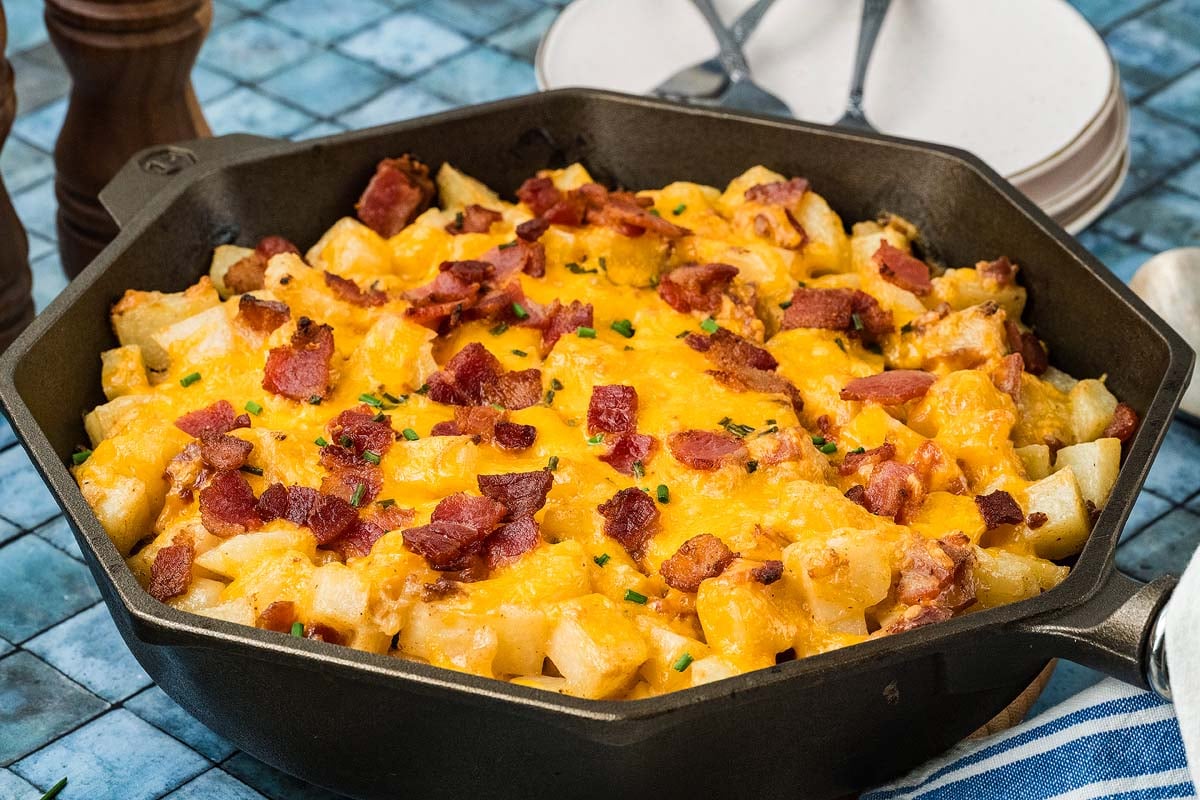 Cast iron skillet filled with diced potatoes topped with melted cheddar cheese and bacon bits.
