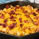 Cast iron skillet filled with diced potatoes topped with melted cheddar cheese, bacon bits, and chives.