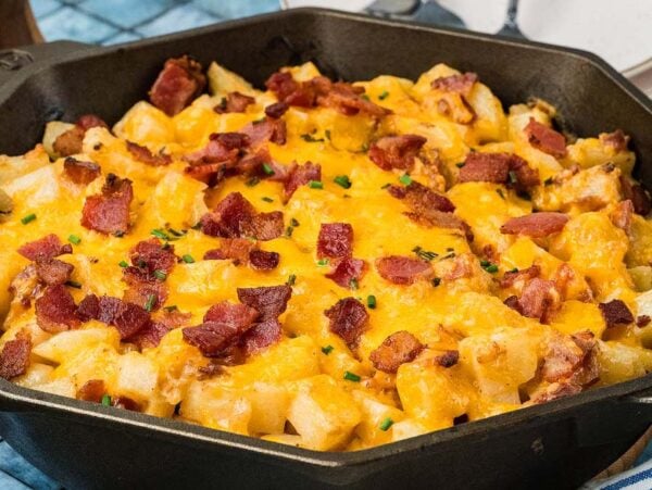 Cast iron skillet filled with diced potatoes topped with melted cheddar cheese, bacon bits, and chives.