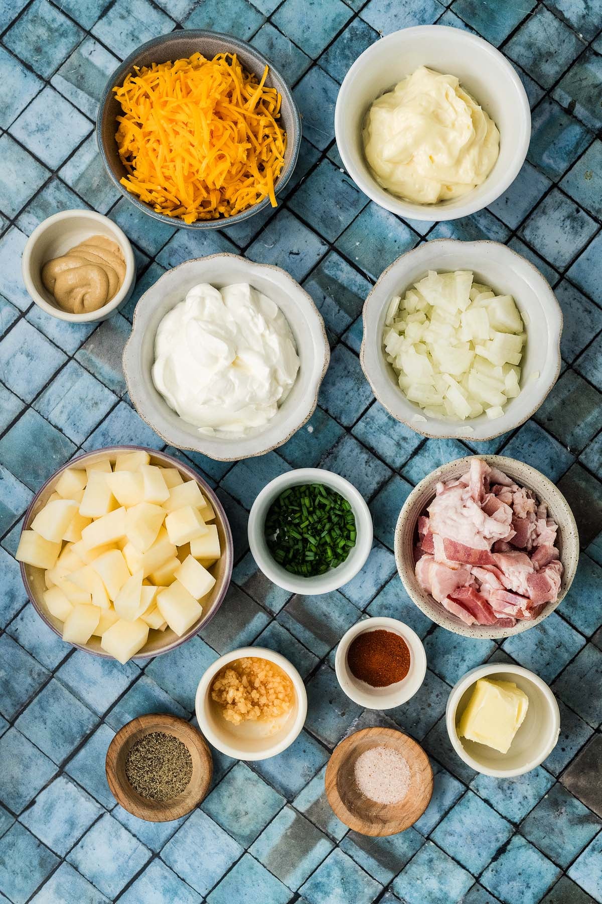 Small bowls containing ingredients for a potato dish, including cubed potatoes, bacon, cheese, sour cream, and spices.