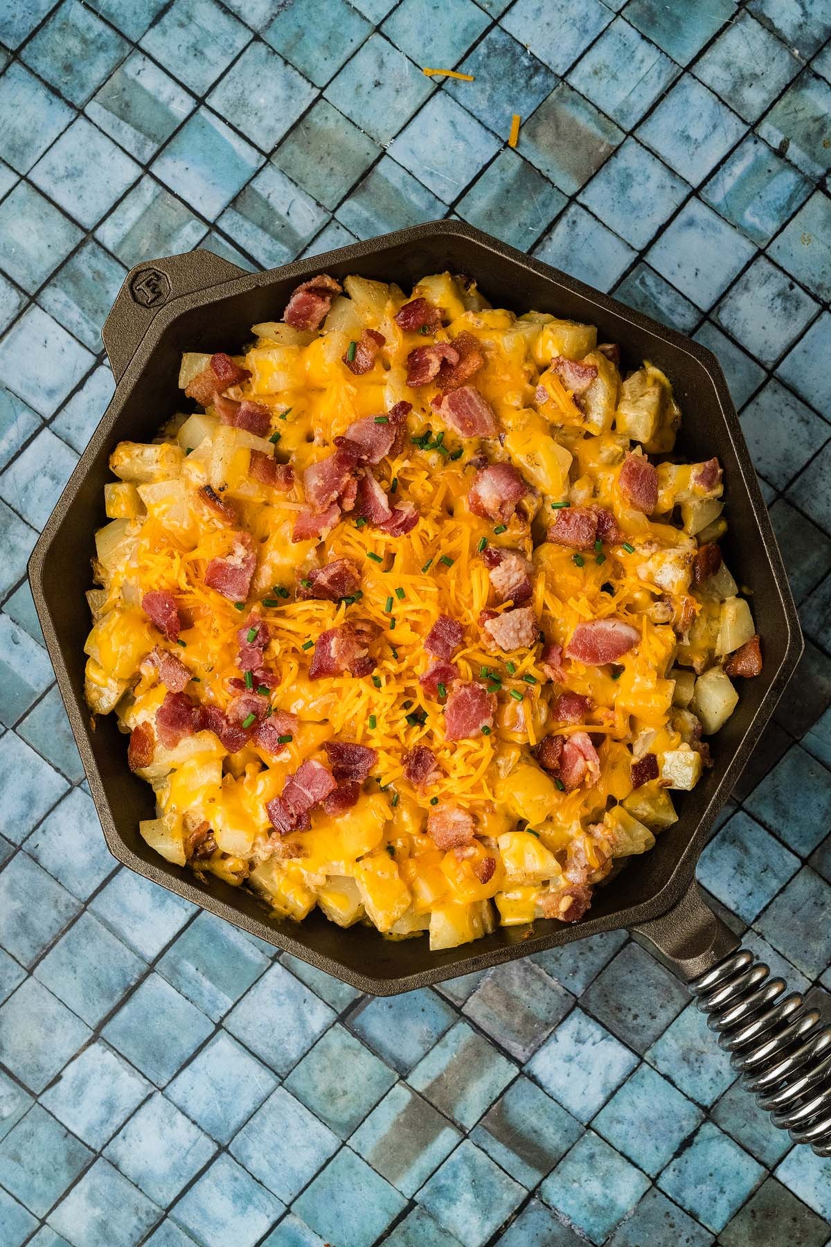 Cast iron skillet filled with diced potatoes topped with melted cheddar cheese, bacon bits, and chives.