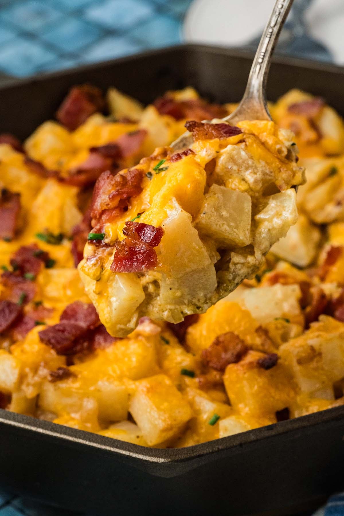 A metal spoon scoops cheesy potatoes topped with bacon from a skillet.