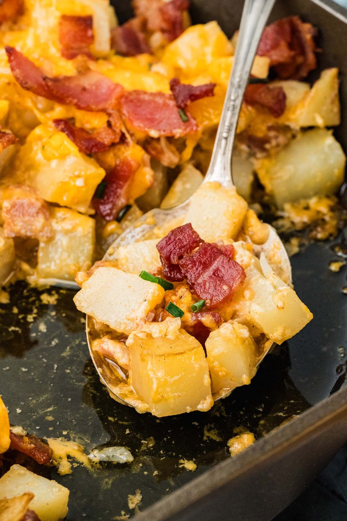 A spoonful of cheesy potatoes with bacon bits held over a baking dish.