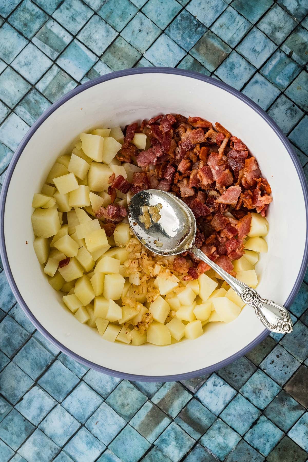 A bowl containing cubed potatoes, chopped bacon, and sautéed onions with a serving spoon.