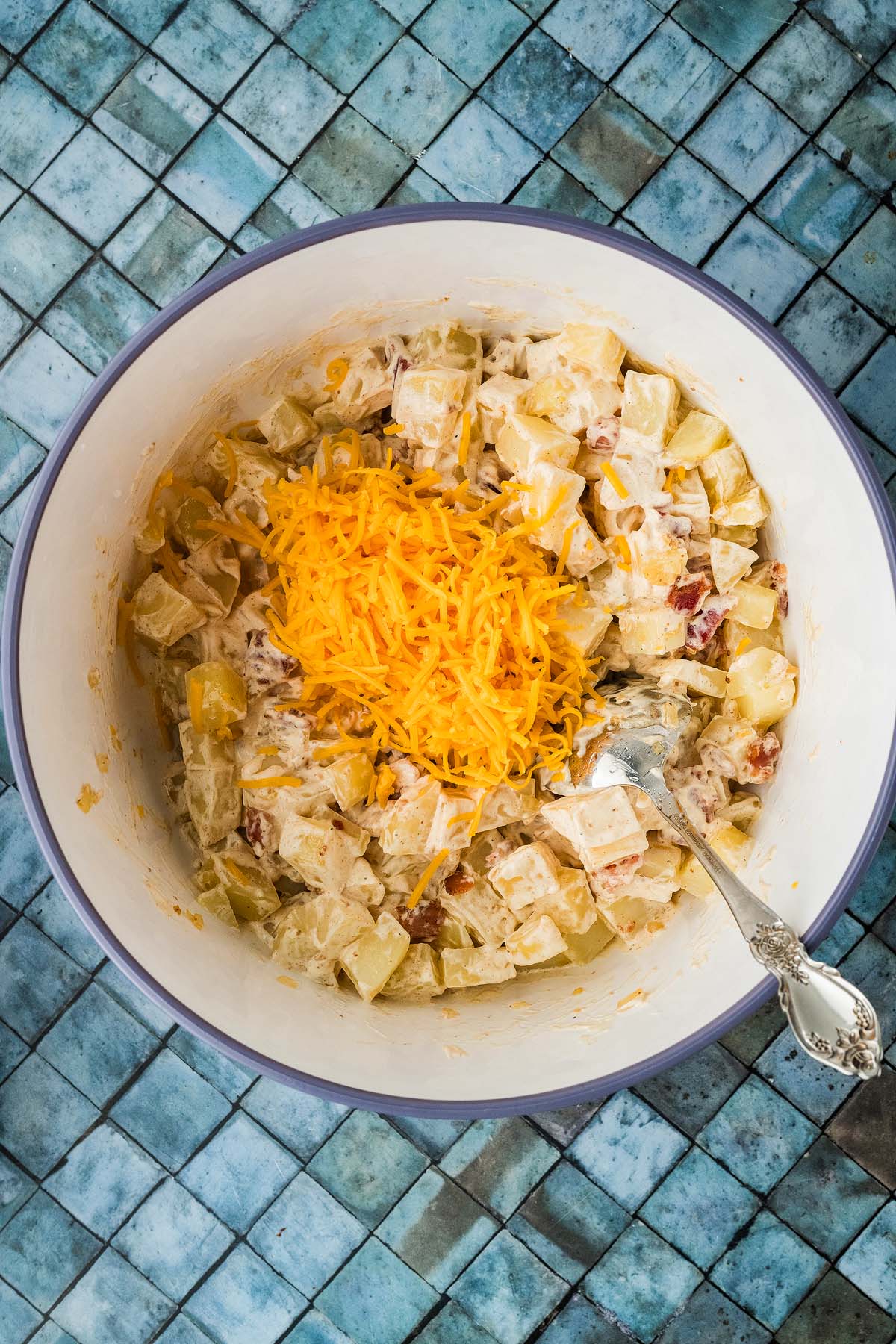 Bowl of cooked potatoes topped with shredded cheddar cheese and a serving spoon.
