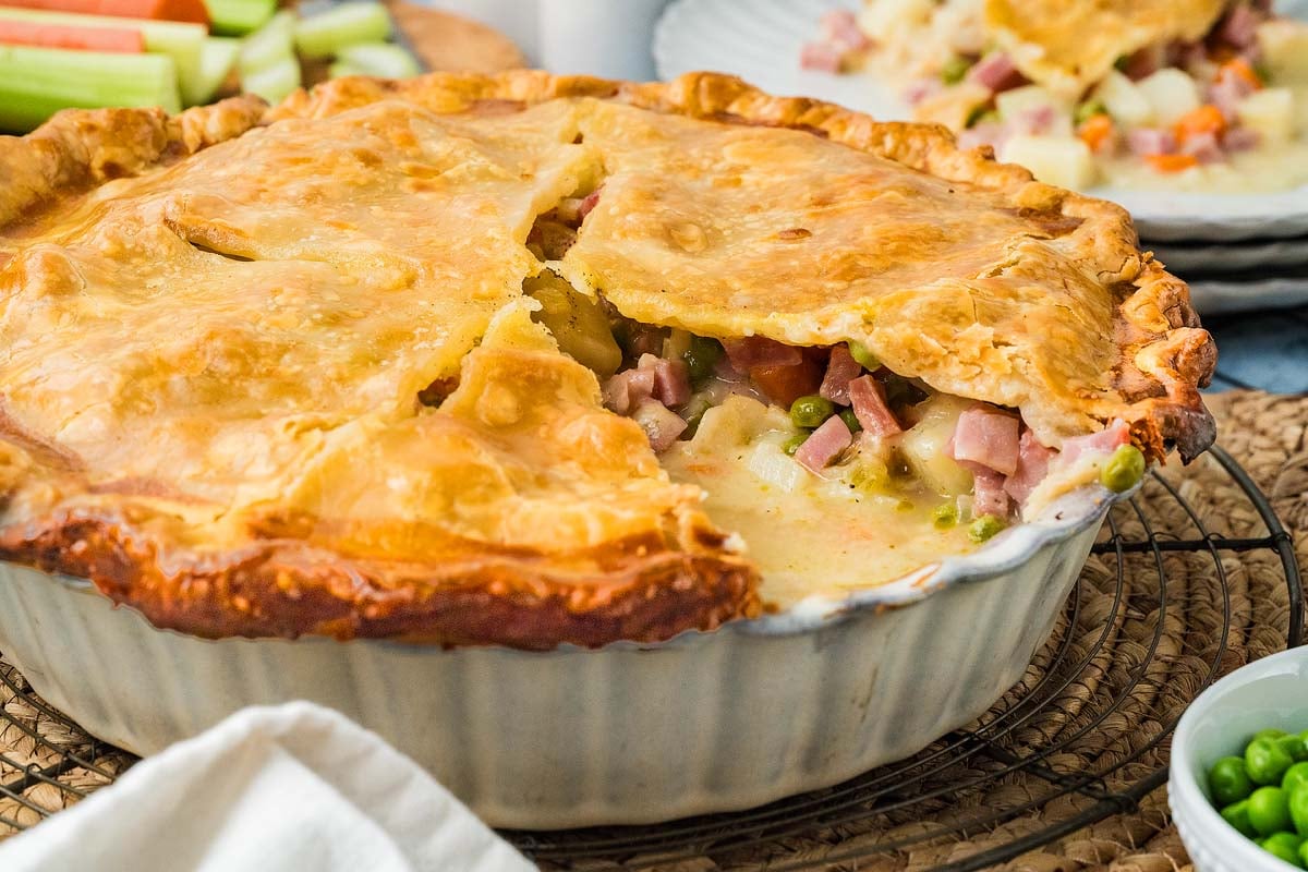A golden-brown ham pot pie with a portion removed to reveal a creamy filling of ham, potatoes, and peas.