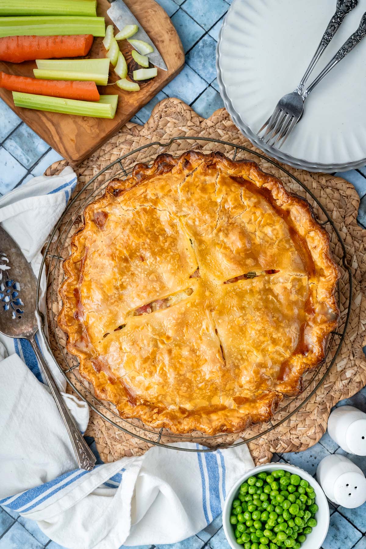 A golden-brown baked pot pie on a wire cooling rack, with celery, carrots, and a bowl of peas nearby.