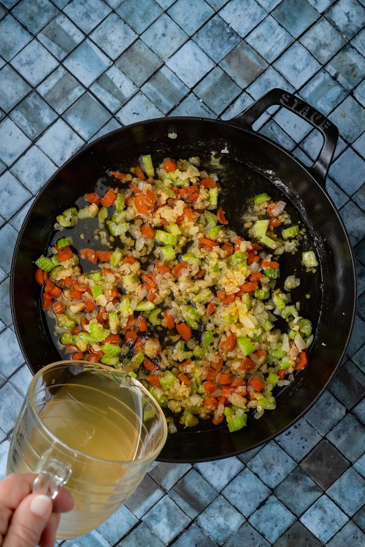 Hand pouring broth into a skillet of sautéed carrots, celery, and onions.