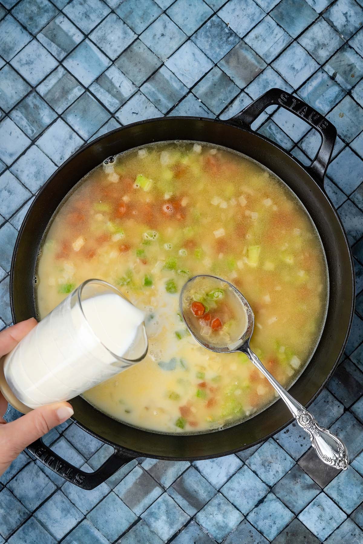 Cream being poured into a cast-iron pot of vegetable soup with a silver spoon nearby.