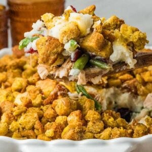 A wooden spoon lifting a scoop of stuffing, turkey, and green bean casserole from a baking dish.
