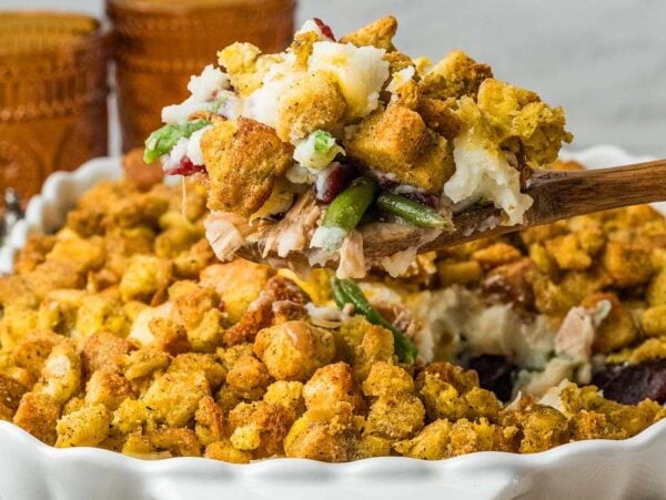 A wooden spoon lifting a scoop of stuffing, turkey, and green bean casserole from a baking dish.