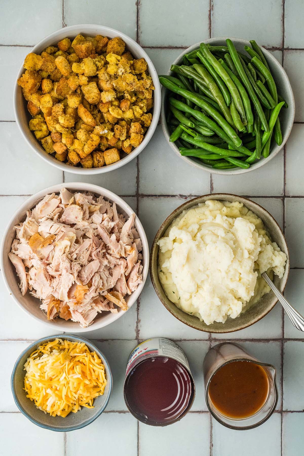 Bowls of shredded turkey, stuffing, green beans, mashed potatoes, shredded cheese, gravy, and a can of cranberries.