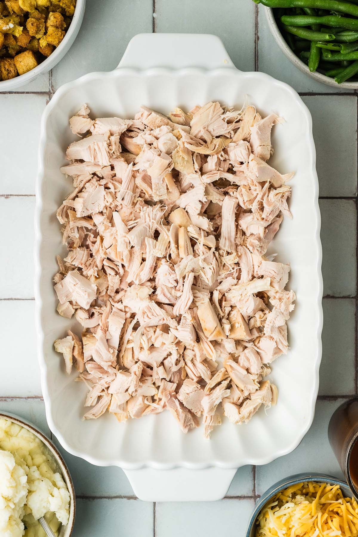 A white baking dish filled with shredded cooked turkey, surrounded by bowls of stuffing, green beans, and potatoes.