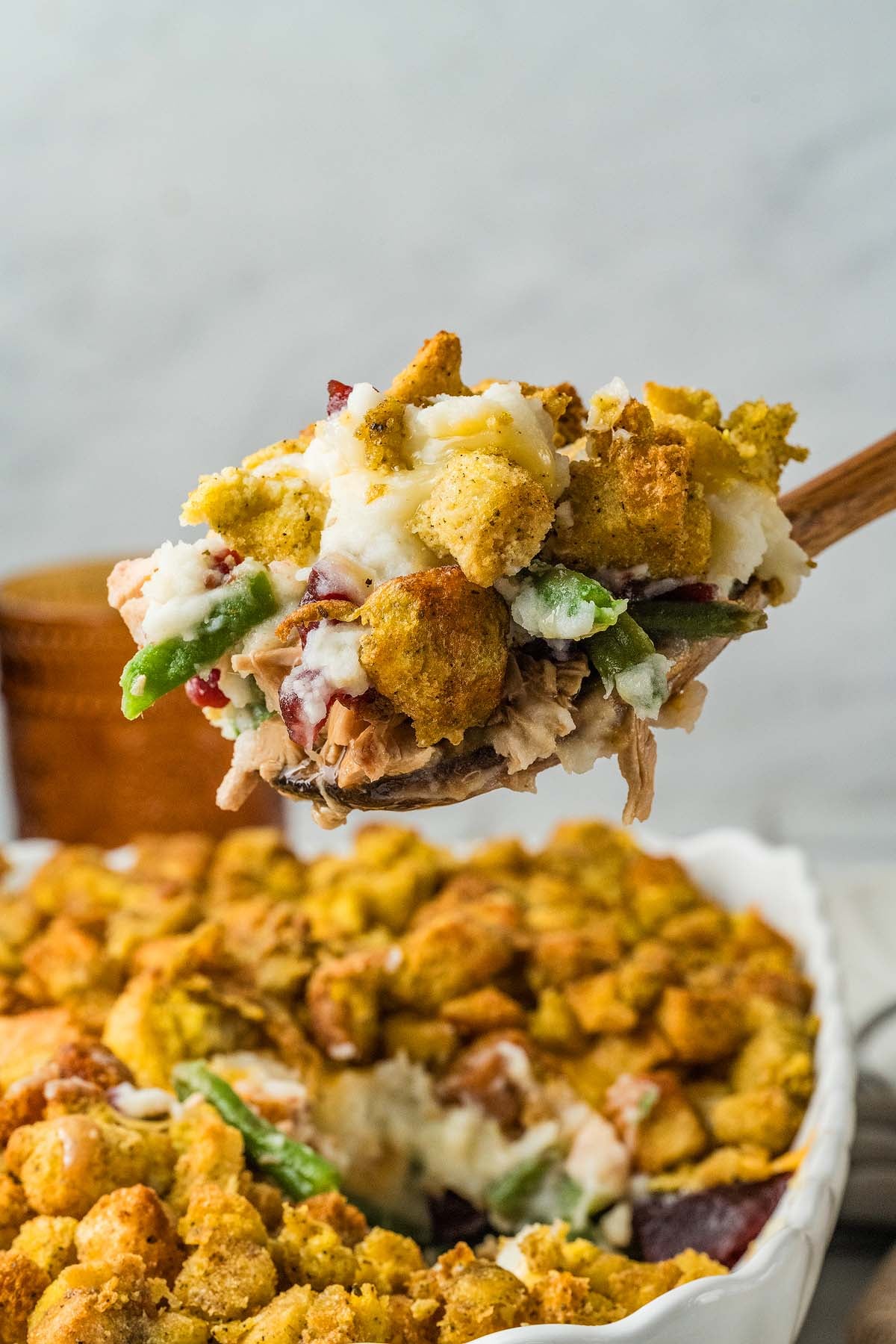 A wooden spoon lifting a scoop of Thanksgiving casserole with turkey, stuffing, mashed potatoes, and green beans.