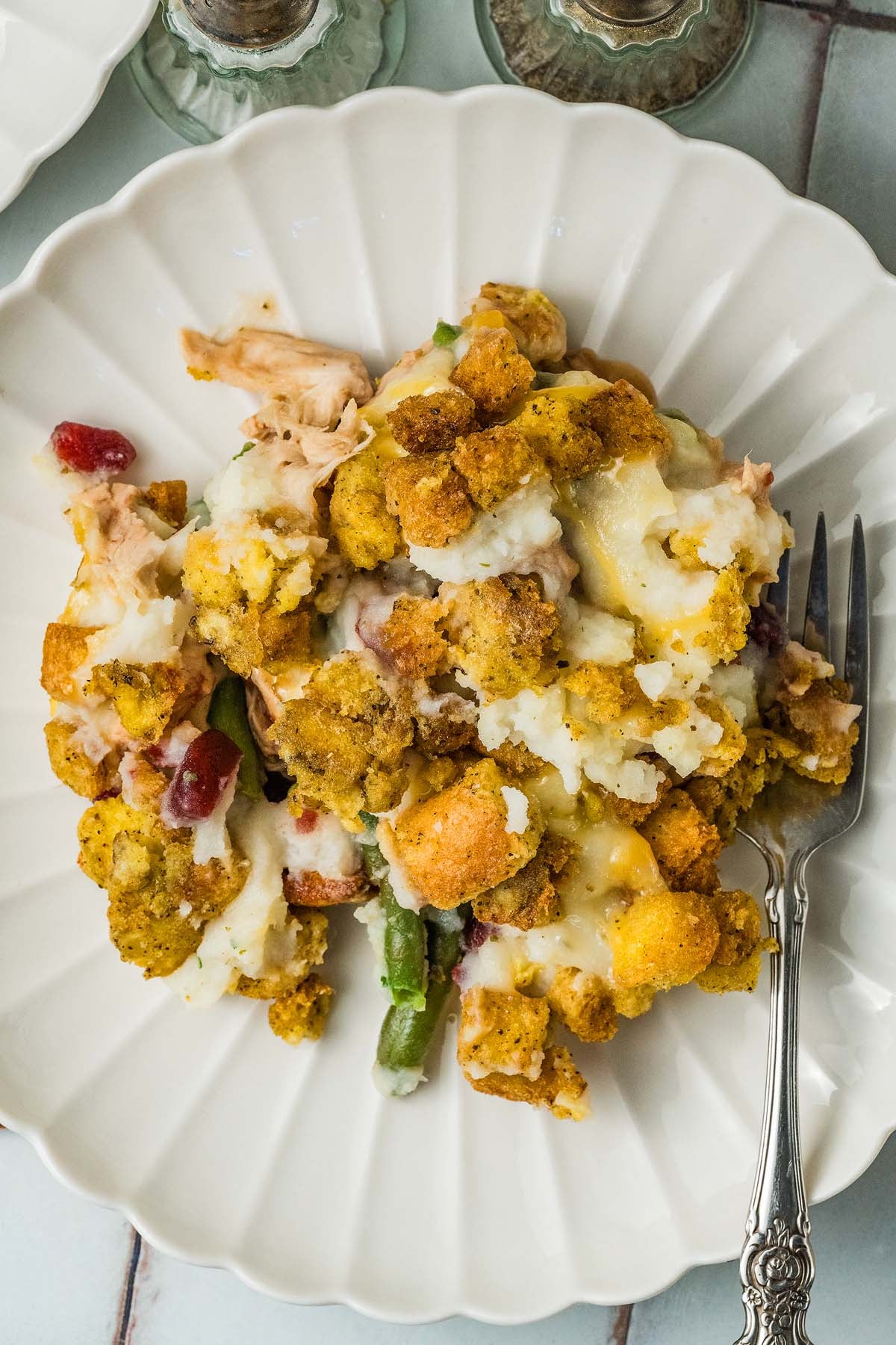 A serving of leftover turkey, mashed potatoes, green beans, and stuffing on a scalloped white plate with a fork.