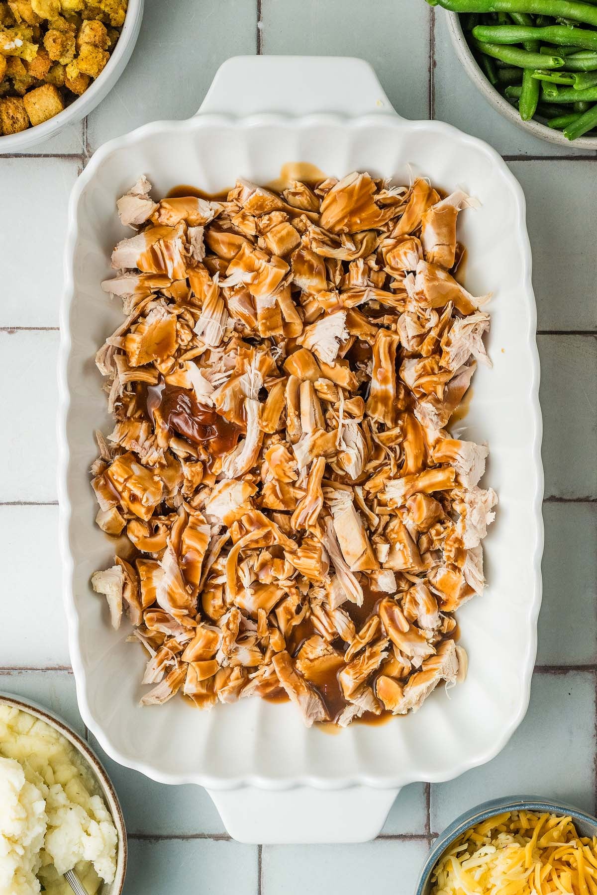 A baking dish of shredded turkey with gravy, surrounded by bowls of stuffing, green beans, mashed potatoes, and cheese.
