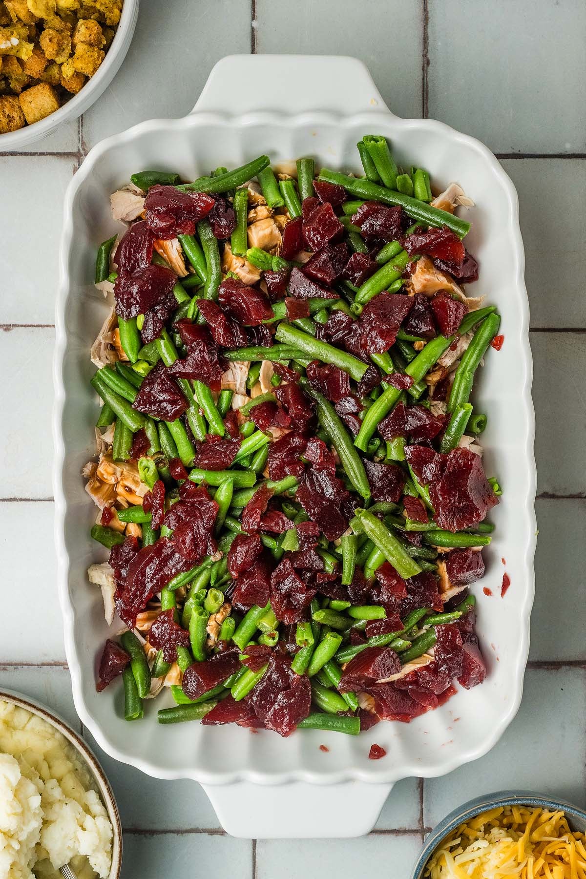 Casserole dish containing turkey, green beans, and cranberry sauce, with bowls of stuffing and mashed potatoes nearby.