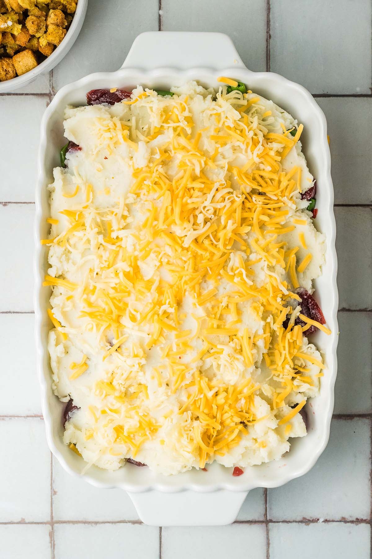 Casserole topped with mashed potatoes and shredded cheddar cheese in a white baking dish.