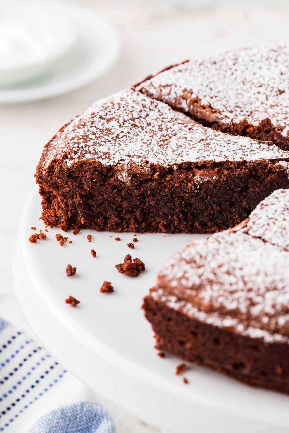A sliced chocolate flourless torte dusted with powdered sugar on a white cake stand.
