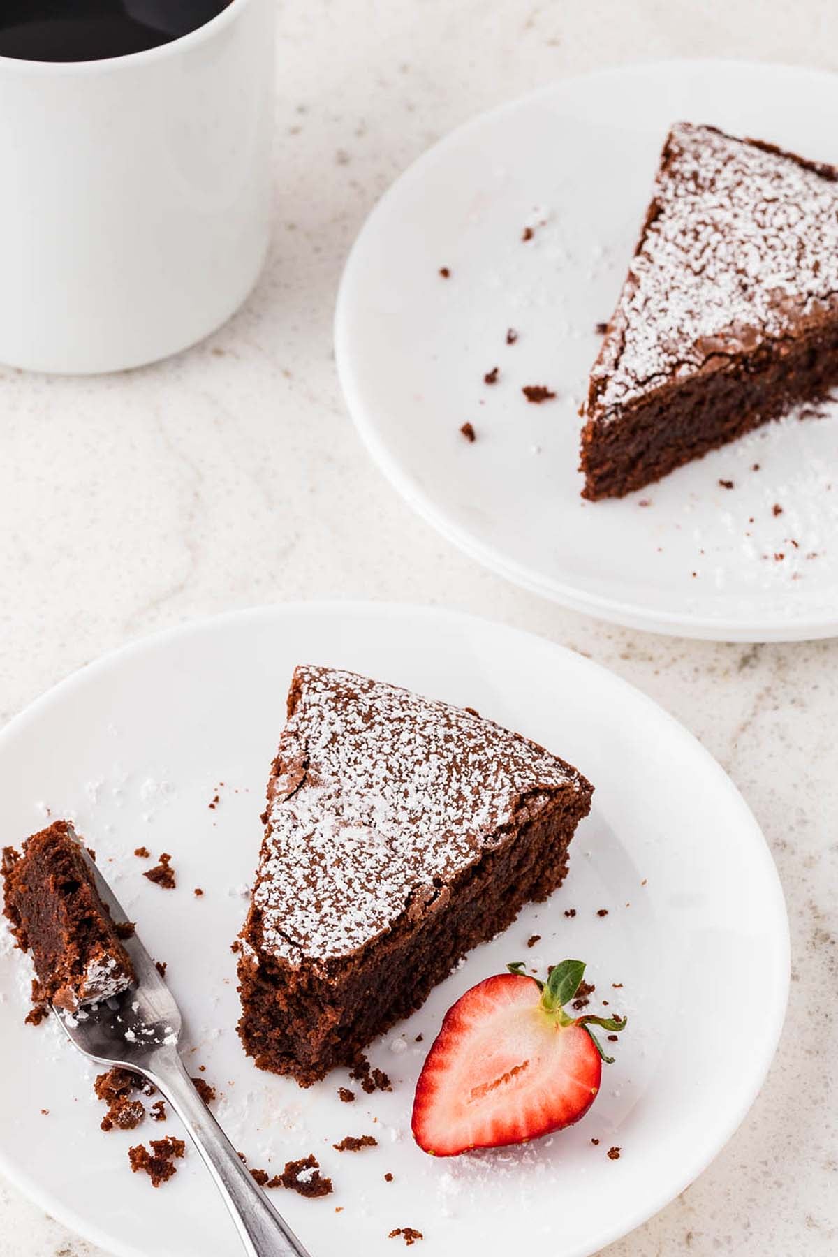 Two plates each holding a slice of chocolate torta caprese cake dusted with powdered sugar, served with a strawberry.