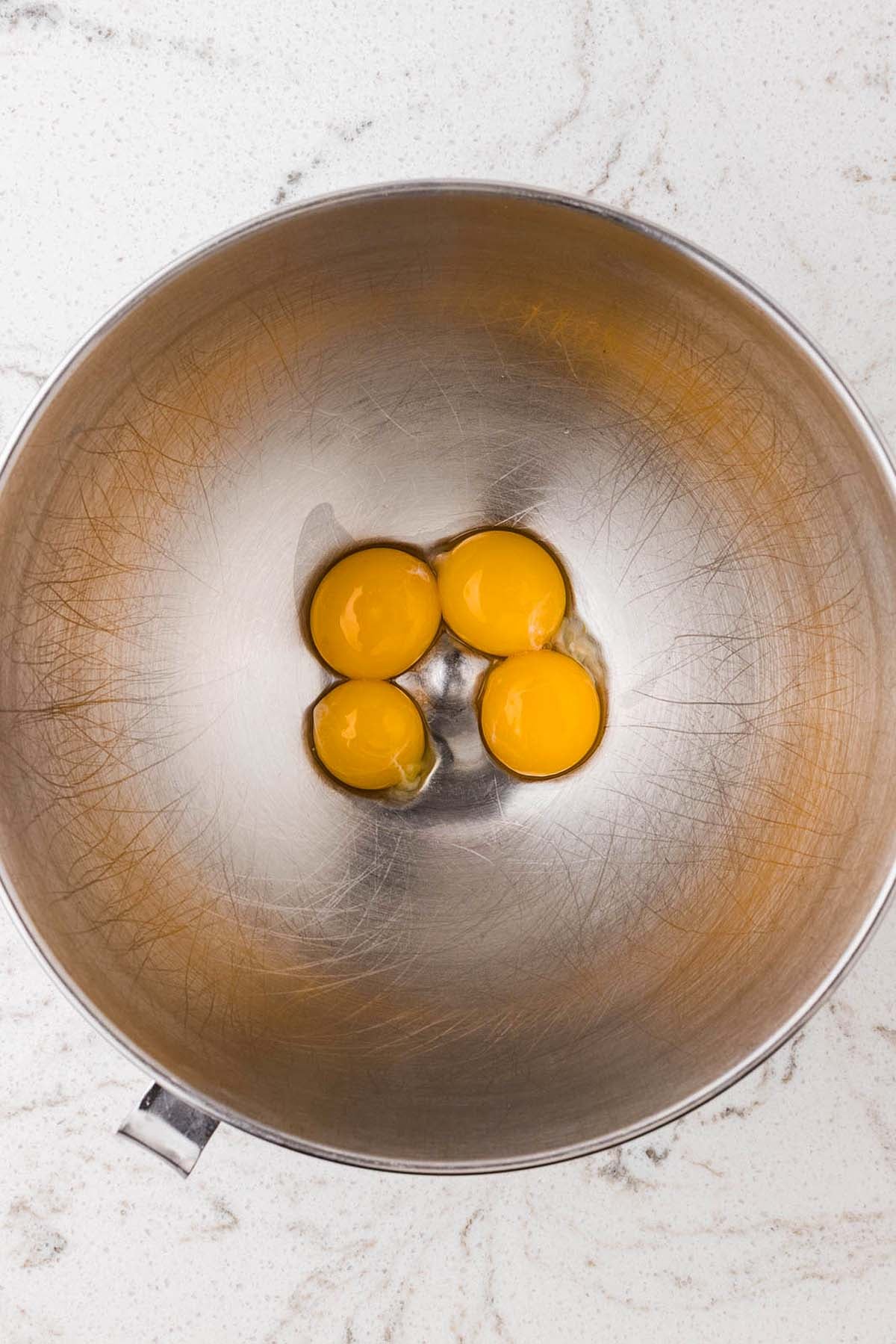 Four raw egg yolks in a stainless steel mixing bowl.