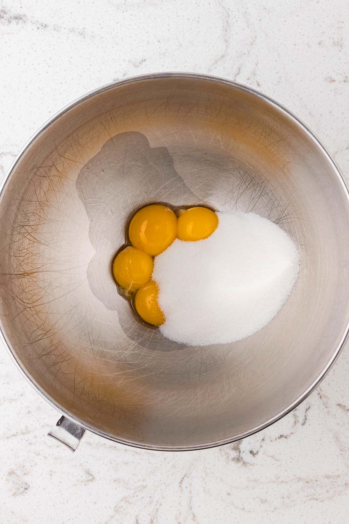 Four egg yolks and a pile of granulated sugar in a metal mixing bowl.