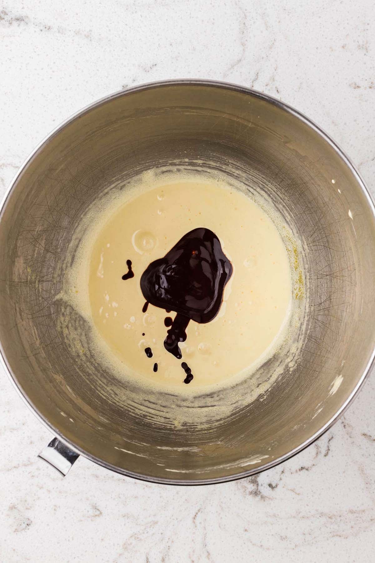 Dark liquid being poured into a pale yellow mixture inside a metal mixing bowl.