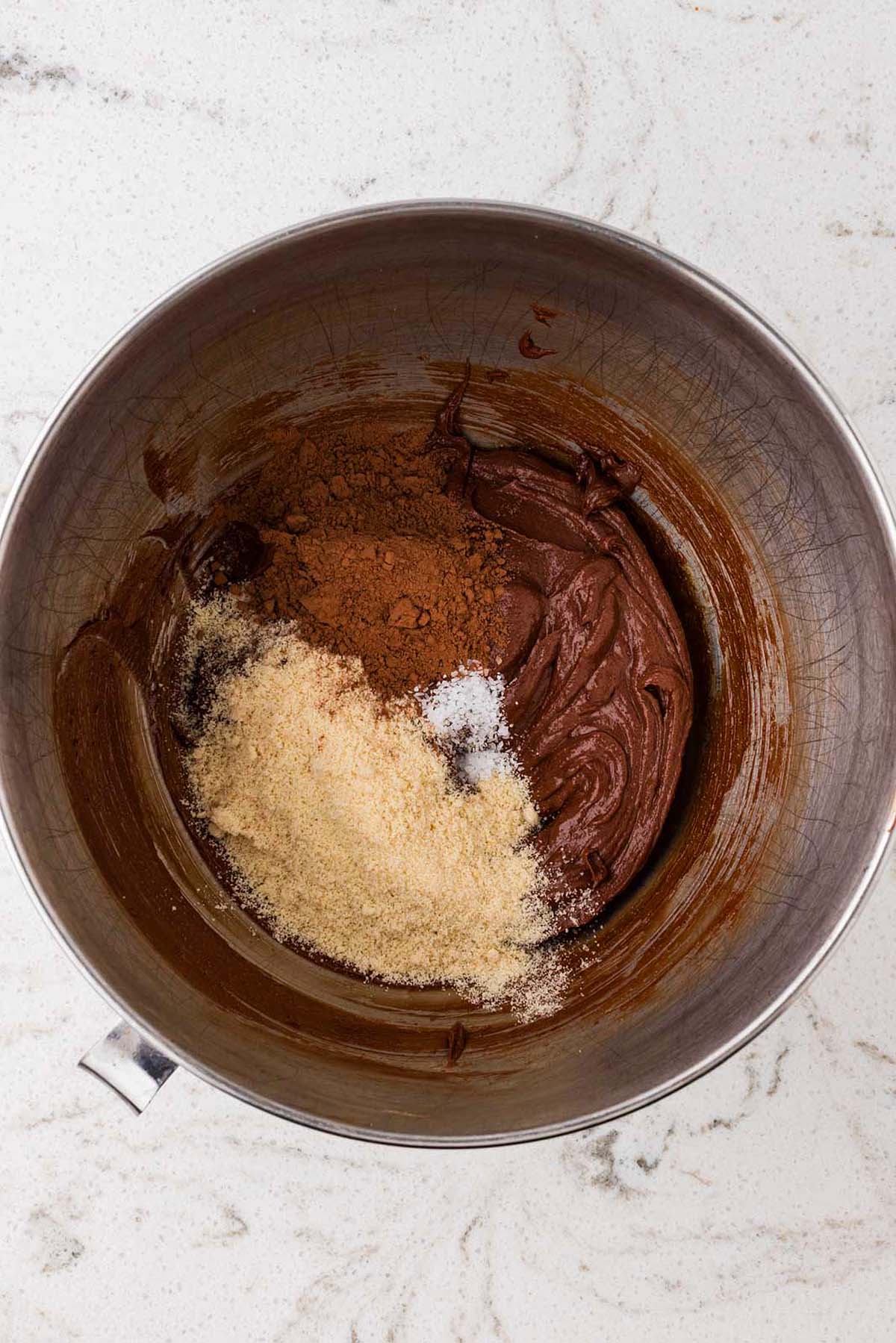 A metal mixing bowl containing chocolate batter with mounds of cocoa powder, granulated sugar, and salt.