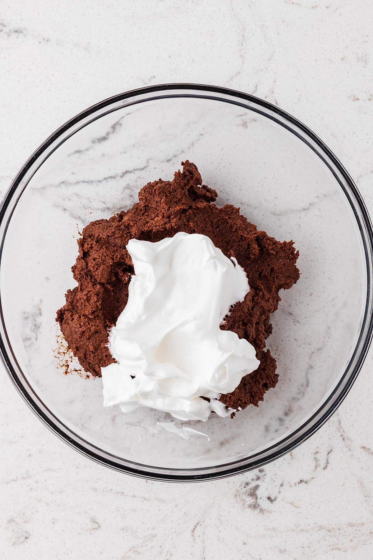 A dollop of white whipped topping sits on top of a mound of dark chocolate batter in a glass mixing bowl.